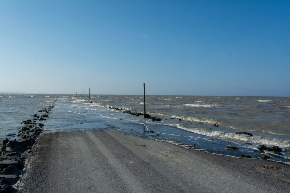 A flooded coastal road surrounded by the sea and horizon under a clear blue sky.